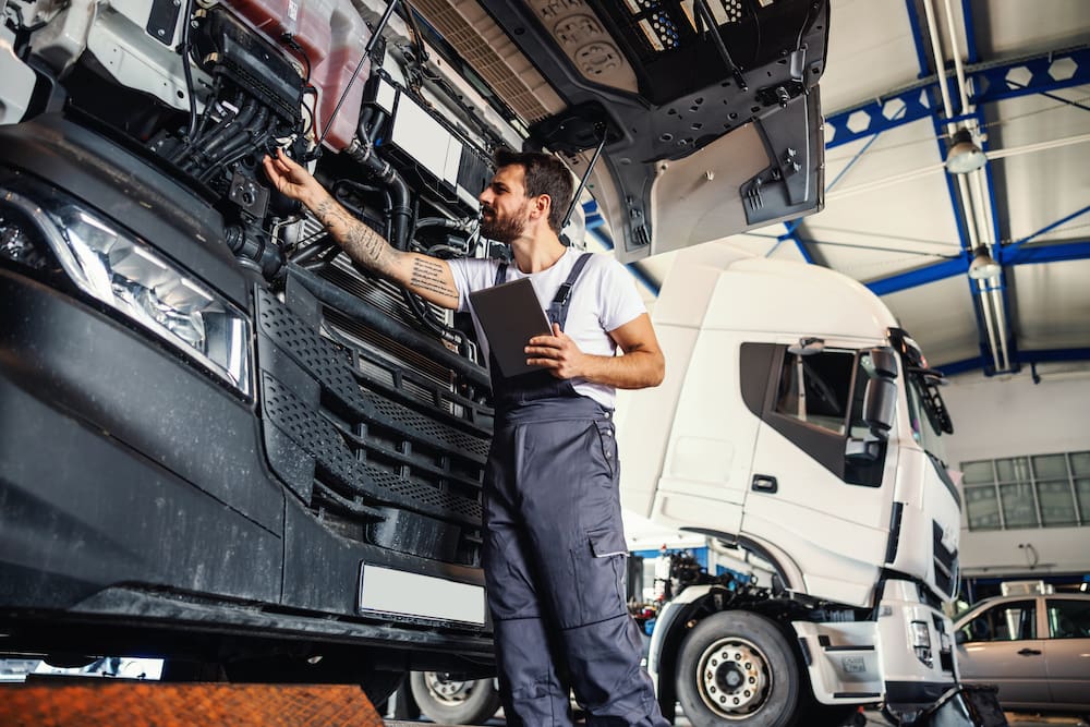 A mechanic inspects the engine of a large commercial truck in a workshop, using the Truckfile app to enhance fleet safety and fleet efficiency. The mechanic, dressed in overalls, holds a tablet to ensure optimal maintenance, with another truck in the background, showcasing the workshop's role in improving fleet performance.