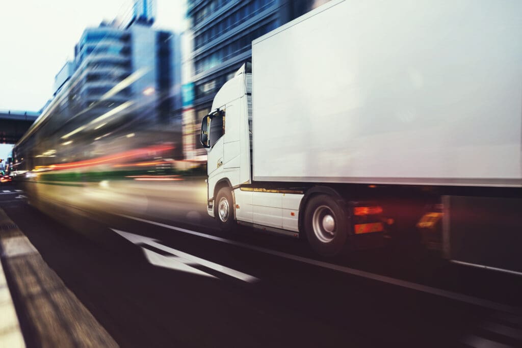 White heavy goods vehicle (HGV) driving through a busy city street, representing urban logistics and the importance of compliance with the Direct Vision Standard (DVS) for enhanced road safety.